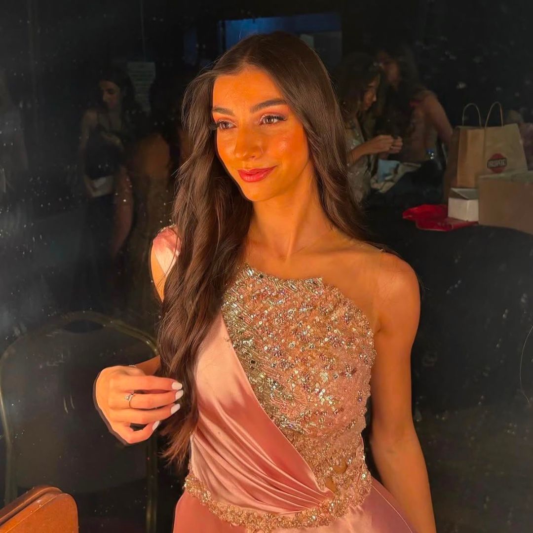 Woman with long brown hair in a pink, sequined one-shoulder dress, smiling softly.