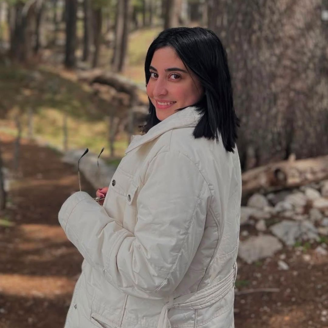 Young woman in white jacket smiles while holding sunglasses in a forested outdoor setting.