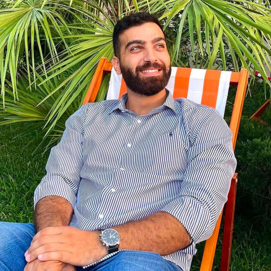 Bearded man in striped shirt and watch sitting in an orange-striped wooden chair outdoors.