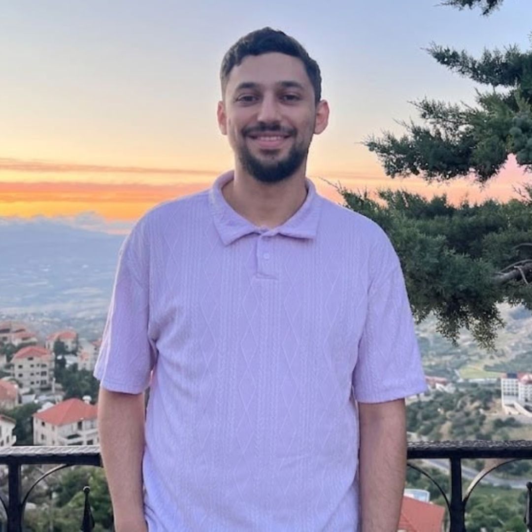 Smiling man with short hair and beard wearing a light purple shirt, sunset and houses in background.