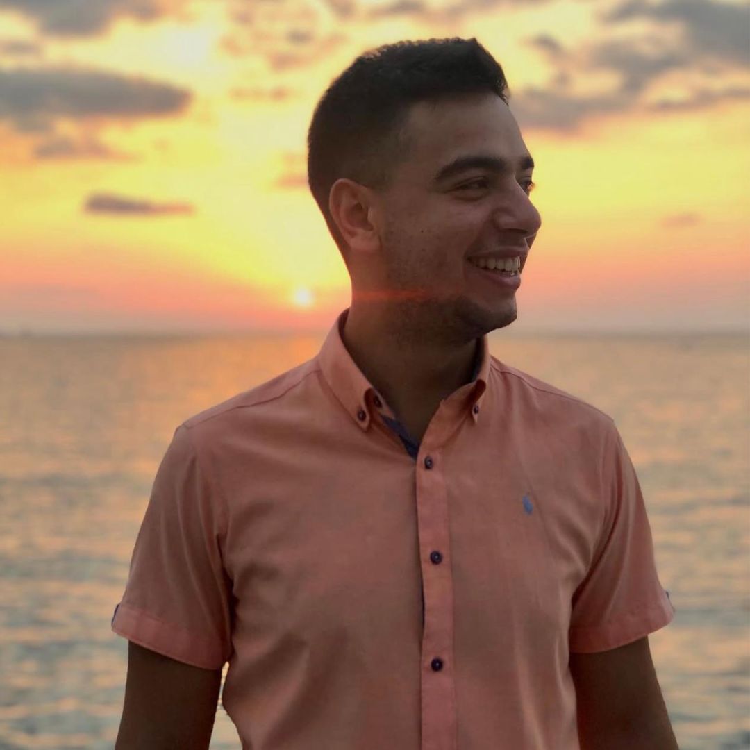 Smiling young man in peach shirt standing by the water at sunset with an orange sky.