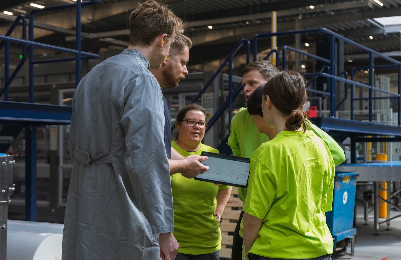 A group of five factory workers, four in bright green shirts, gather around a tablet. 