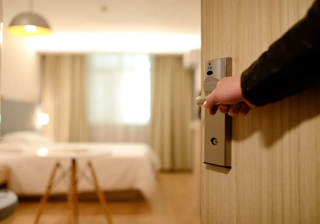 Mano abriendo una puerta de madera equipada con una cerradura electrónica inteligente, con un dormitorio de alojamiento turístico difuminado al fondo