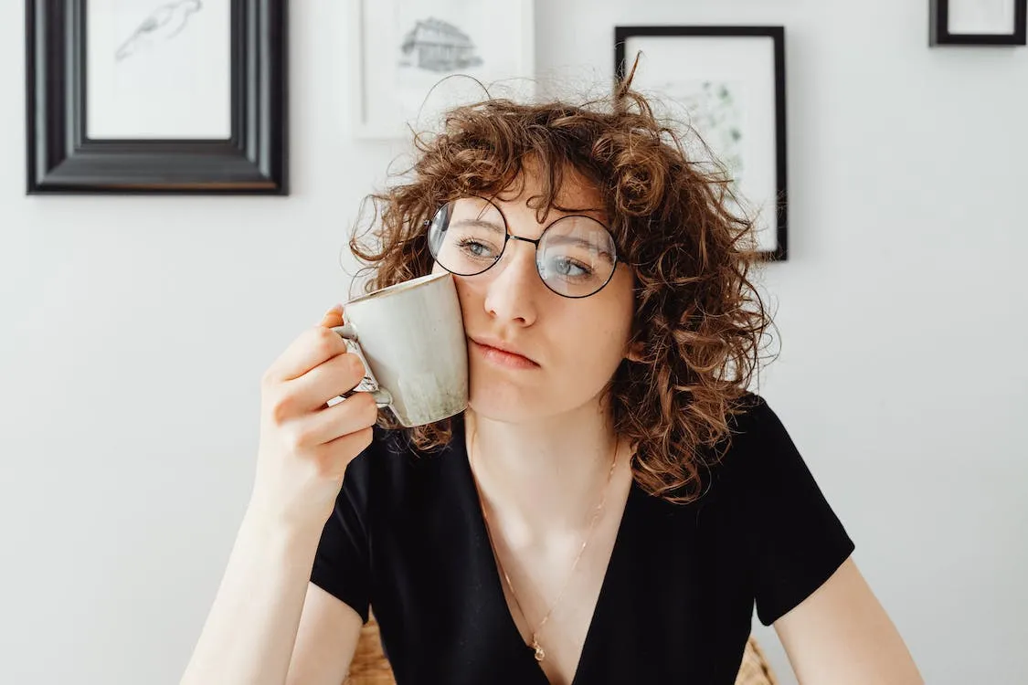 Una mujer con gafas redondas y pelo rizado sostiene una taza contra su mejilla con expresión de preocupación, reflejando la inquietud de un propietario ante problemas con inquilinos