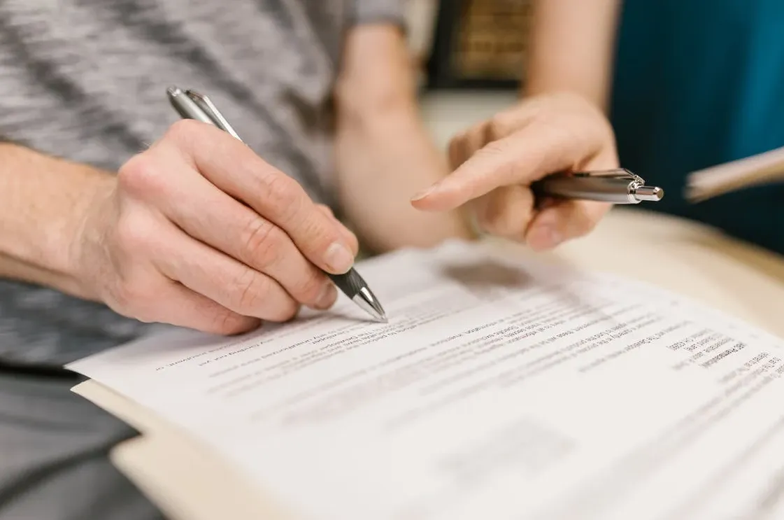 Primer plano de una mano firmando un documento en papel con un bolígrafo mientras otra persona señala el texto con el dedo, representando la firma de un contrato de arras