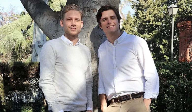 Dos hombres jóvenes vestidos con camisas blancas y ropa casual sonríen junto al tronco de un gran árbol en un parque, ilustrando la entrevista a los fundadores de Box2box