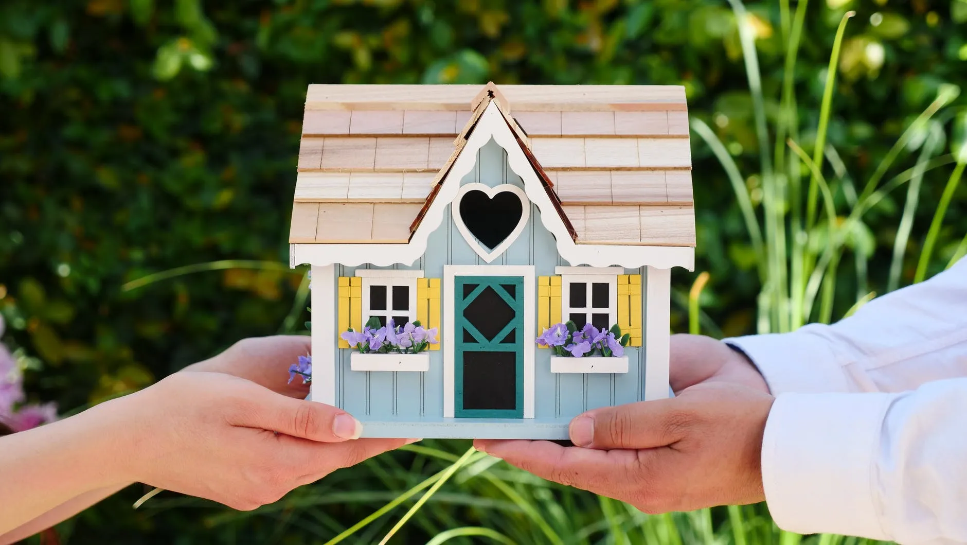 Dos pares de manos sostienen una maqueta de casa de madera azul claro con un corazón calado y flores en las ventanas sobre un fondo verde de jardín, simbolizando la adquisición de una hipoteca verde