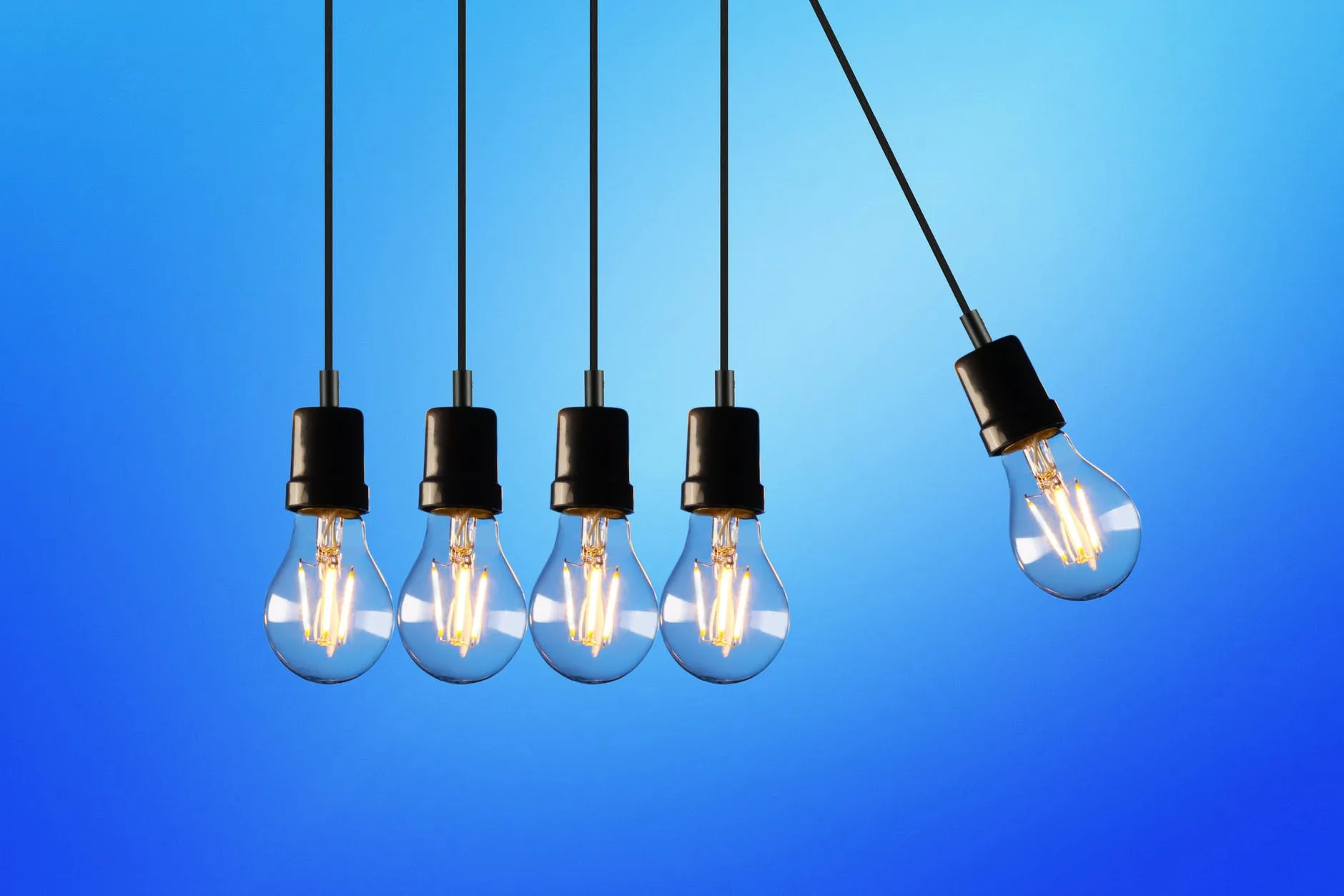 Cinco bombillas encendidas con filamentos brillantes cuelgan sobre un fondo azul, dispuestas como un péndulo de Newton con la bombilla derecha oscilando hacia arriba, ilustrando el ahorro en las tarifas de luz