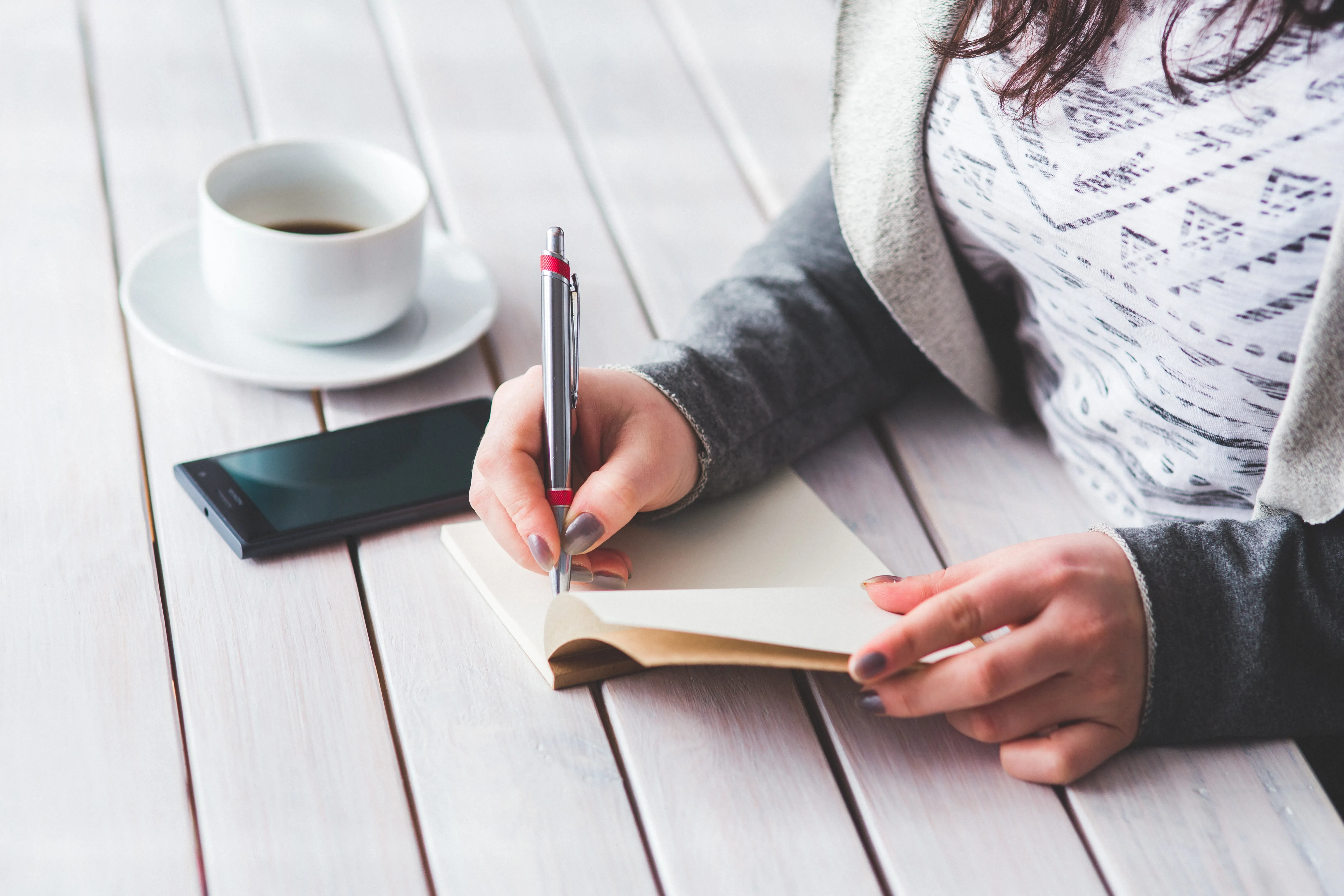 Una persona escribe con un bolígrafo plateado en un cuaderno sobre una mesa de madera clara junto a un teléfono móvil y una taza de café, representando la preparación de un checklist de mudanza