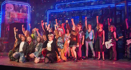 Actors dressed in 80's attire singing with their left arms raised.