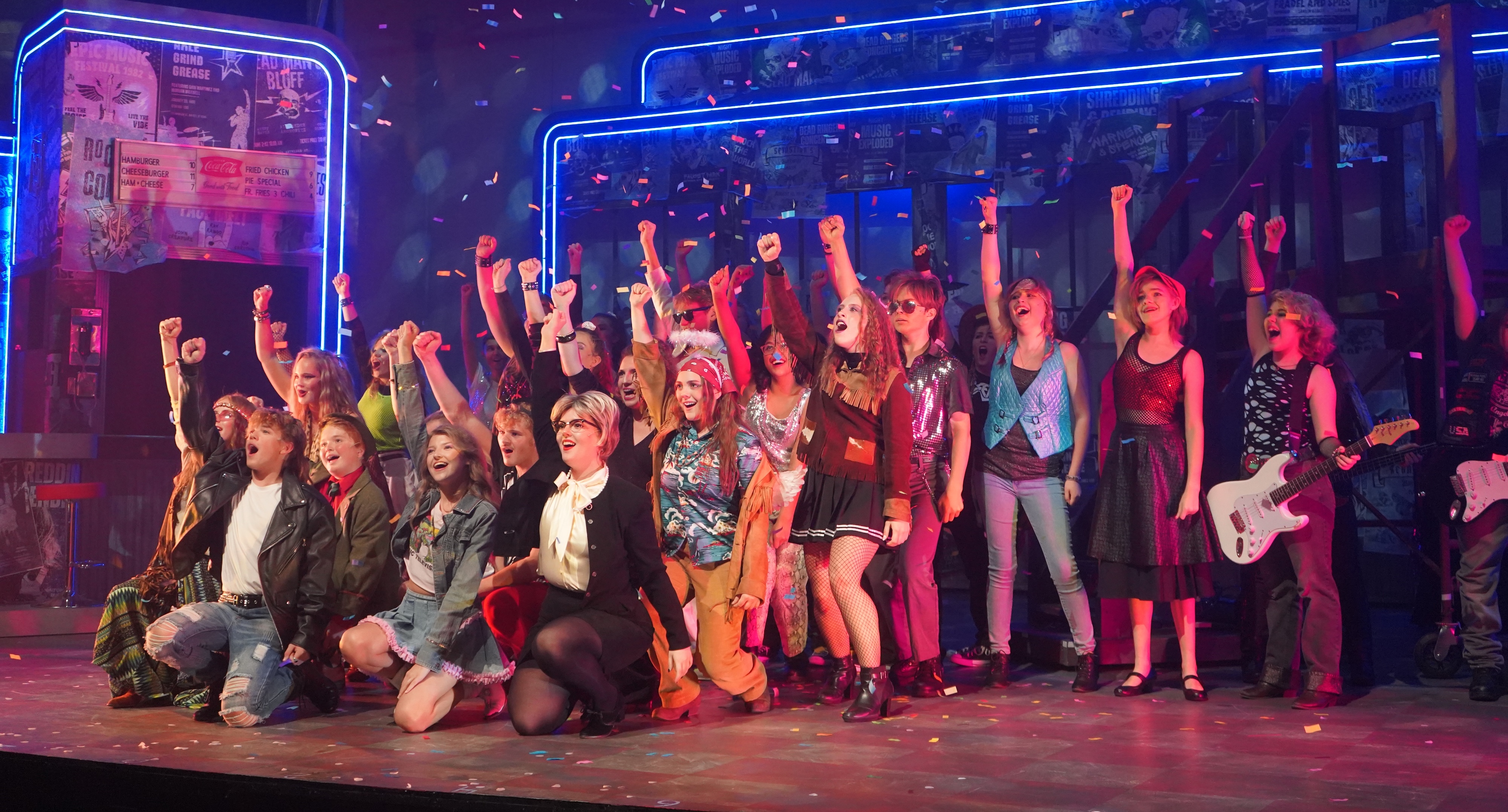 Actors dressed in 80's attire singing with their left arms raised.