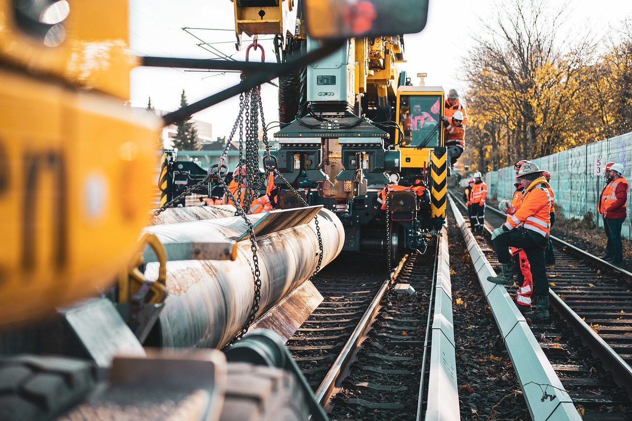 Workers transporting construction materials with heavy machinery over train tracks.