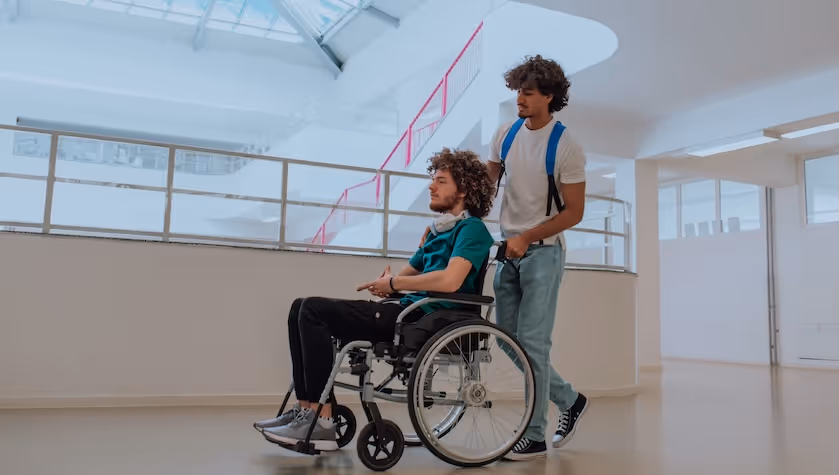 Zwei junge Männer befinden sich in einem Museum. Einer von ihnen ist im Rollstuhl, seine Begleitperson schiebt den Rollstuhl.