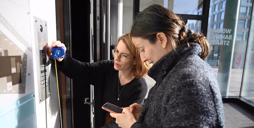 Daniela Schöb von Sitios und Judith Graser von physiopaed erfassen die Zugänglichkeitsinformationen einer Physiopraxis. Sie vermessen die Höhe der Bedienelemente bei der Eingangstüre.