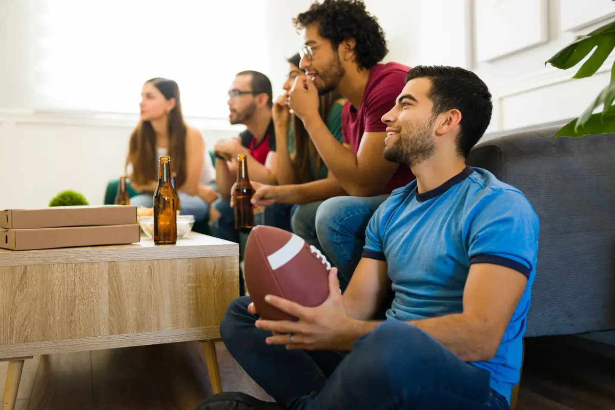 Fantasy Football friends sitting on a couch with drinks and pizza watching a game while holding a football.