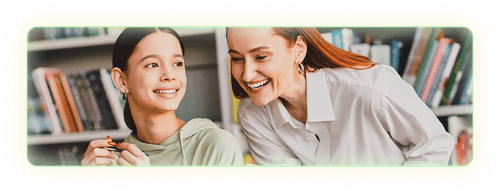 Smiling female teacher leaning over a smiling student's desk to view her work.
