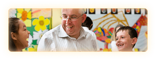 Male teacher smiling as he sits between two elementary aged children in a classroom setting.