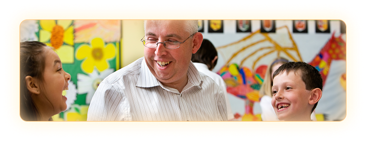 Male teacher smiling as he sits between two elementary aged children in a classroom setting.