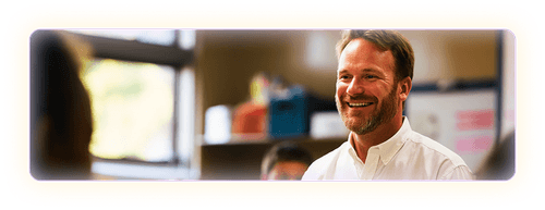 Male teacher in white button up shirt smiling in a classroom setting.