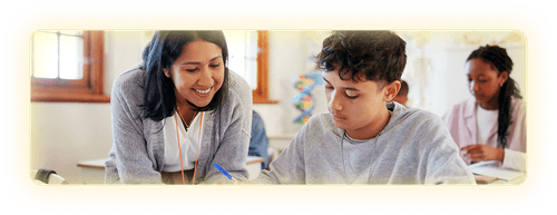 Female teacher leaning over a desk helping a teenage student in a classroom setting.