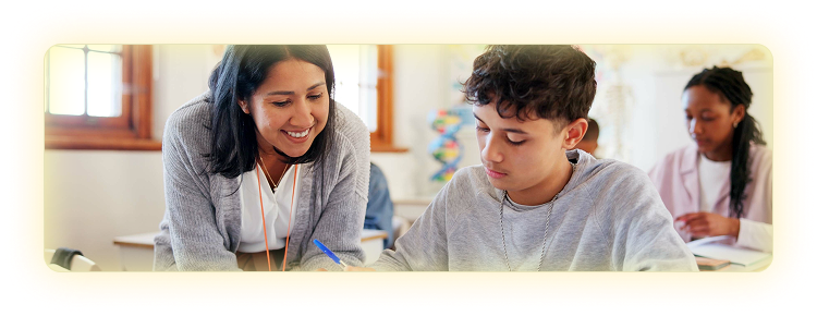 Female teacher leaning over a desk helping a teenage student in a classroom setting.