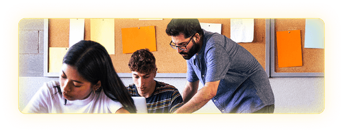 Male teacher leaning over a student's desk to help in in a classroom setting with a female student working at another desk in the foreground.