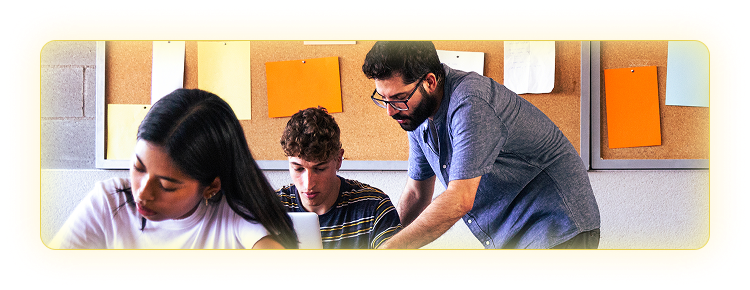 Male teacher leaning over a student's desk to help in in a classroom setting with a female student working at another desk in the foreground. 