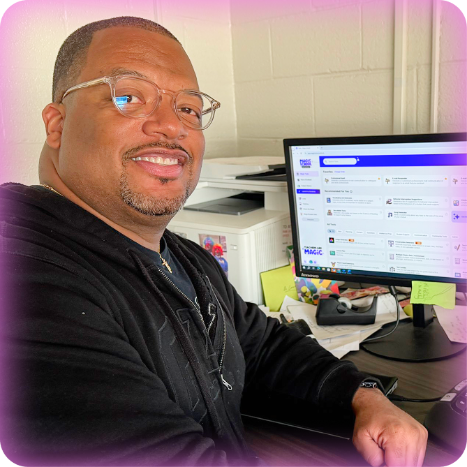 Image of a smiling male teacher sitting at a desk in front a desktop computer with the MagicSchool platform displayed on screen.