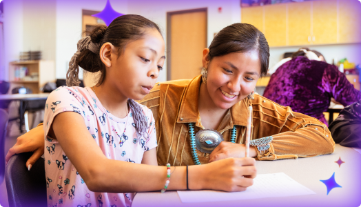 A teacher smiles while working side-by-side with a young student on a writing activity in the classroom.