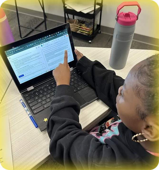 Student using a laptop in a classroom and pointing to the screen where they're using MagicSchool.