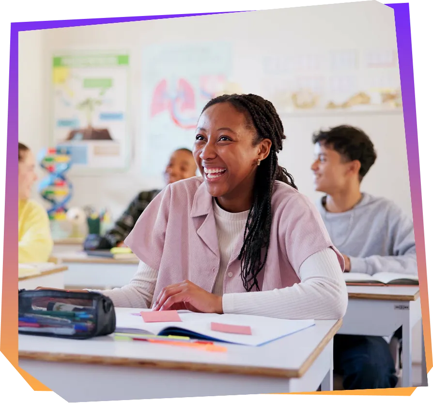 Image of a student sitting at a desk in a classroom and smiling with other students in the background.