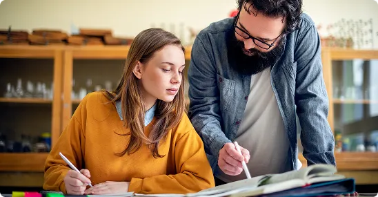 Female student sitting at a desk and writing while a teacher looks over her work and guides her.