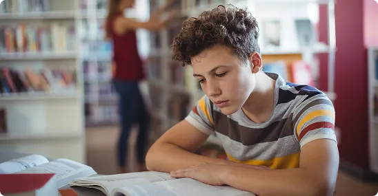 Male student sitting at a table reading a book in a library setting.