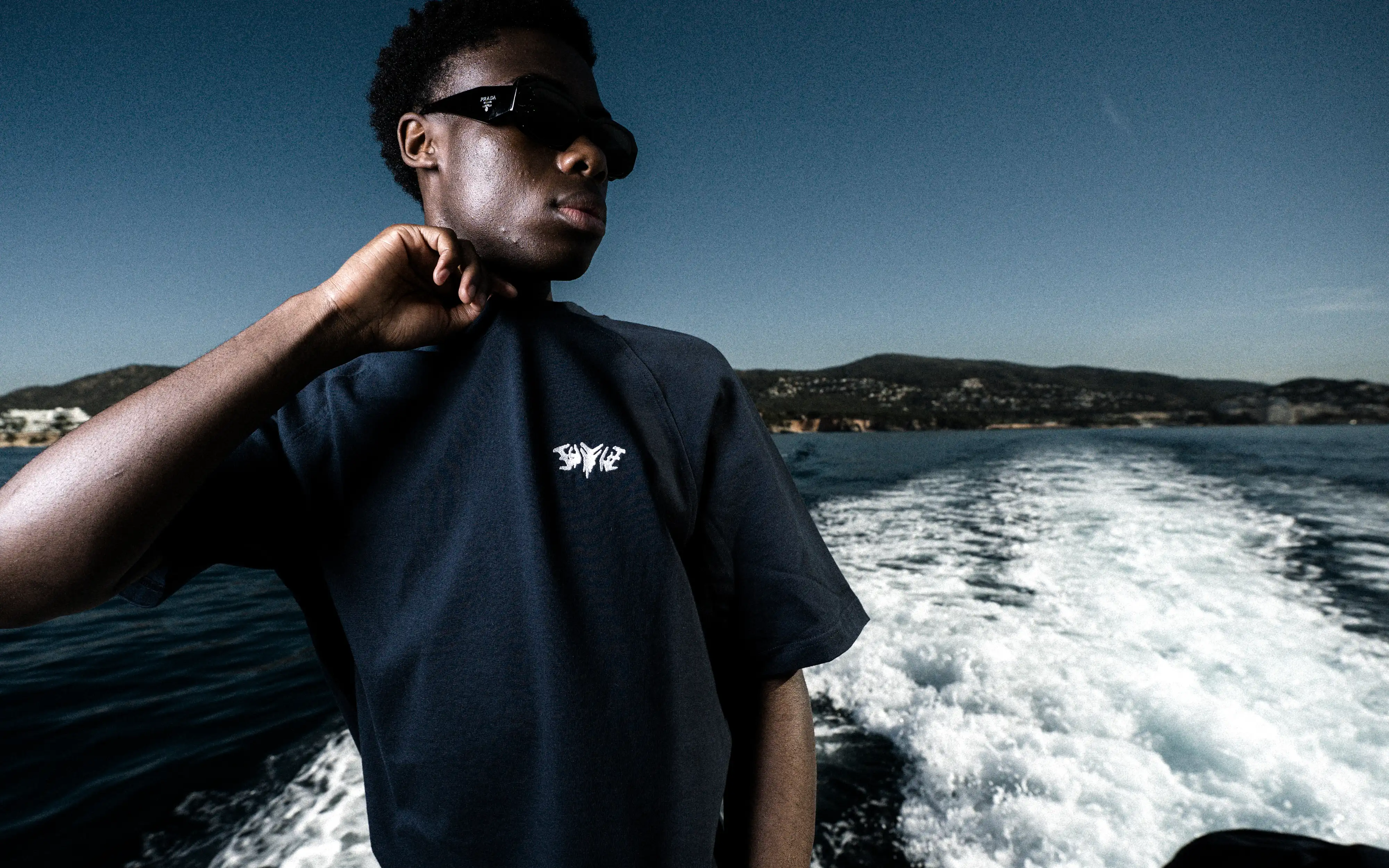Model mit Sonnenbrille im dunkelblauen 45Keys-T-Shirt vor der Heckwelle einer fahrenden Yacht.