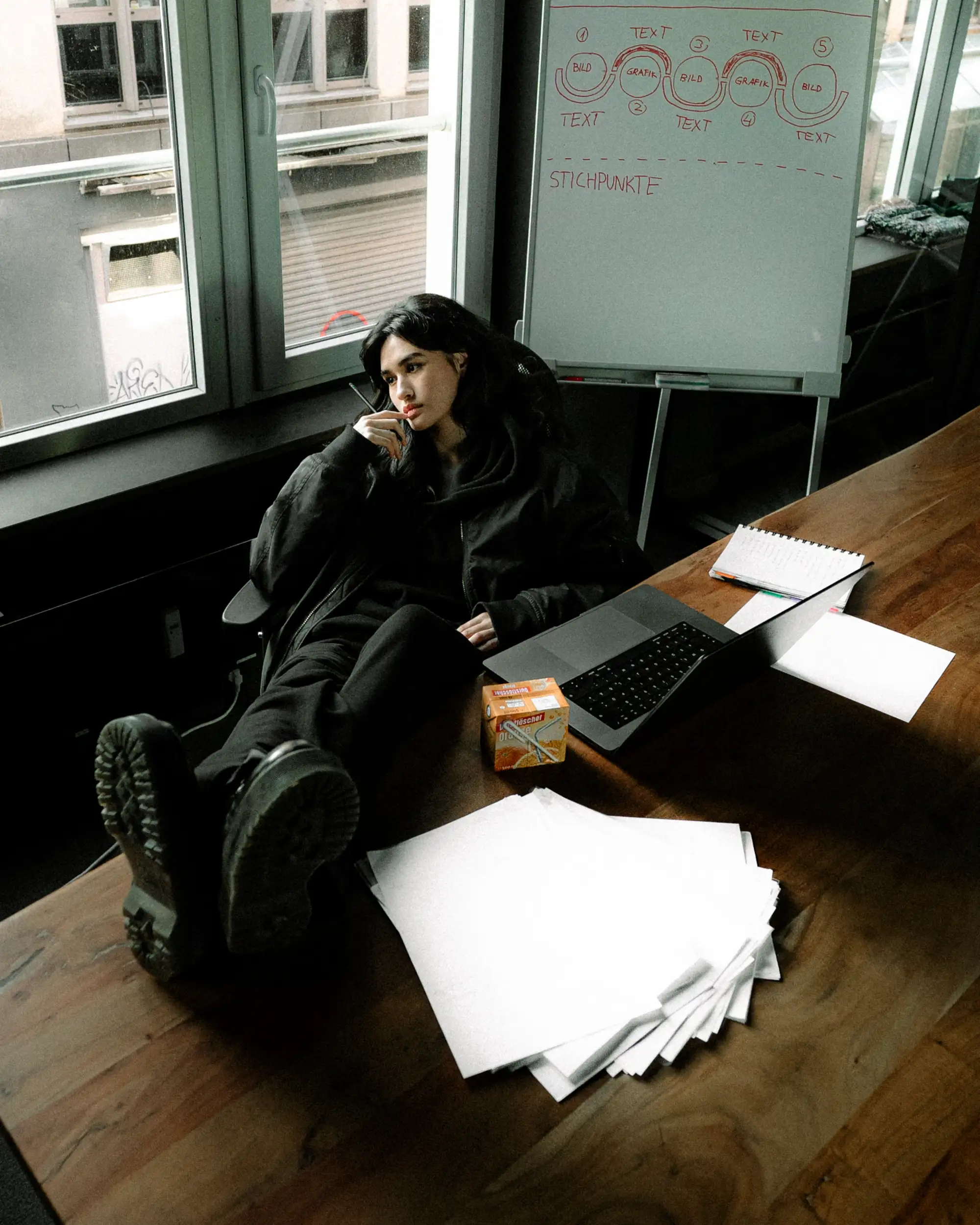 Augustina sitzt in schwarzen Sweatpants auf einem Schreibtisch neben Unterlagen im Wuppertaler Büro.