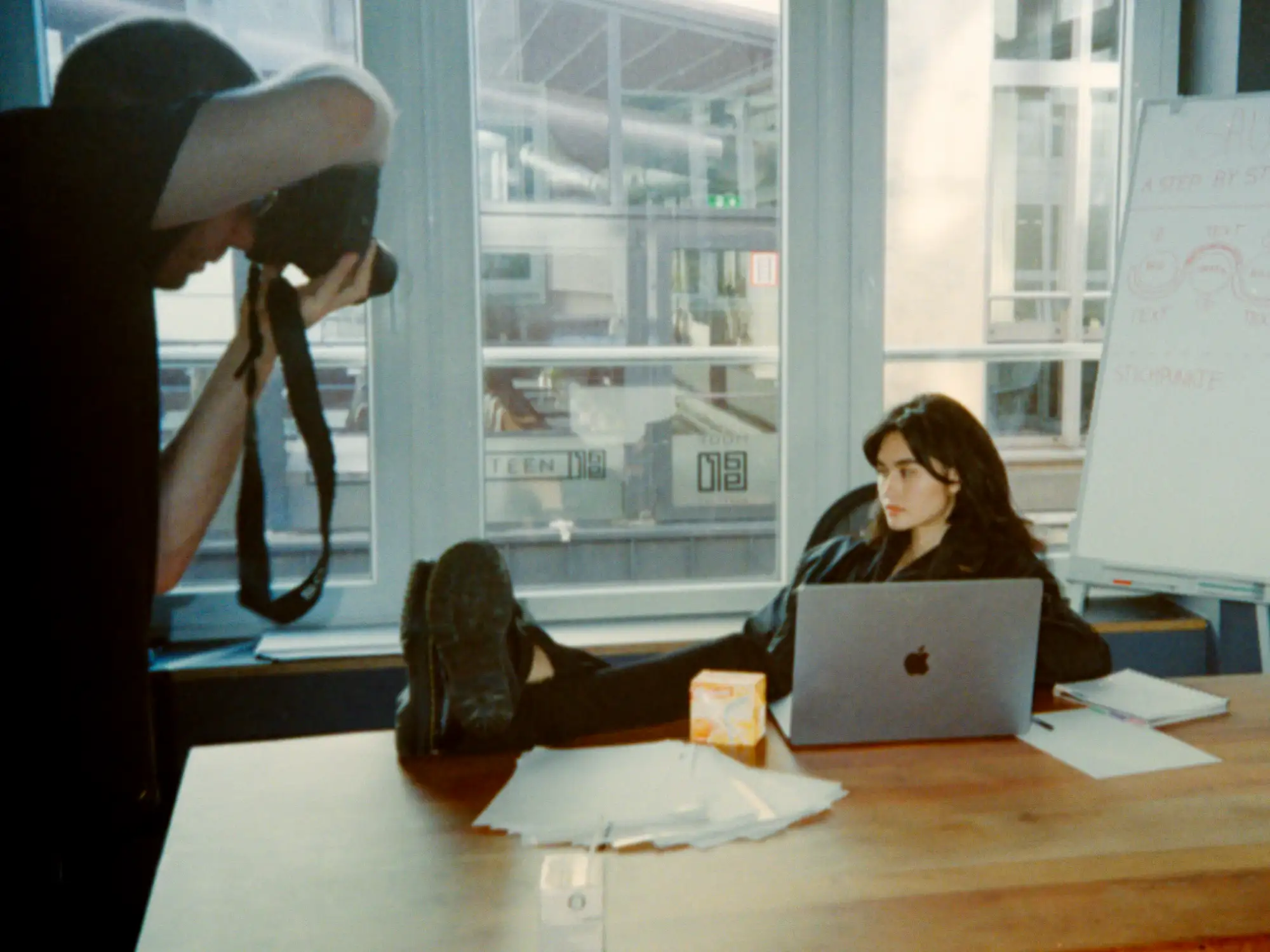 Behind the Scenes: Augustina sitzt hinter einem Laptop am Schreibtisch, während ein Fotograf die Aufnahme vorbereitet.