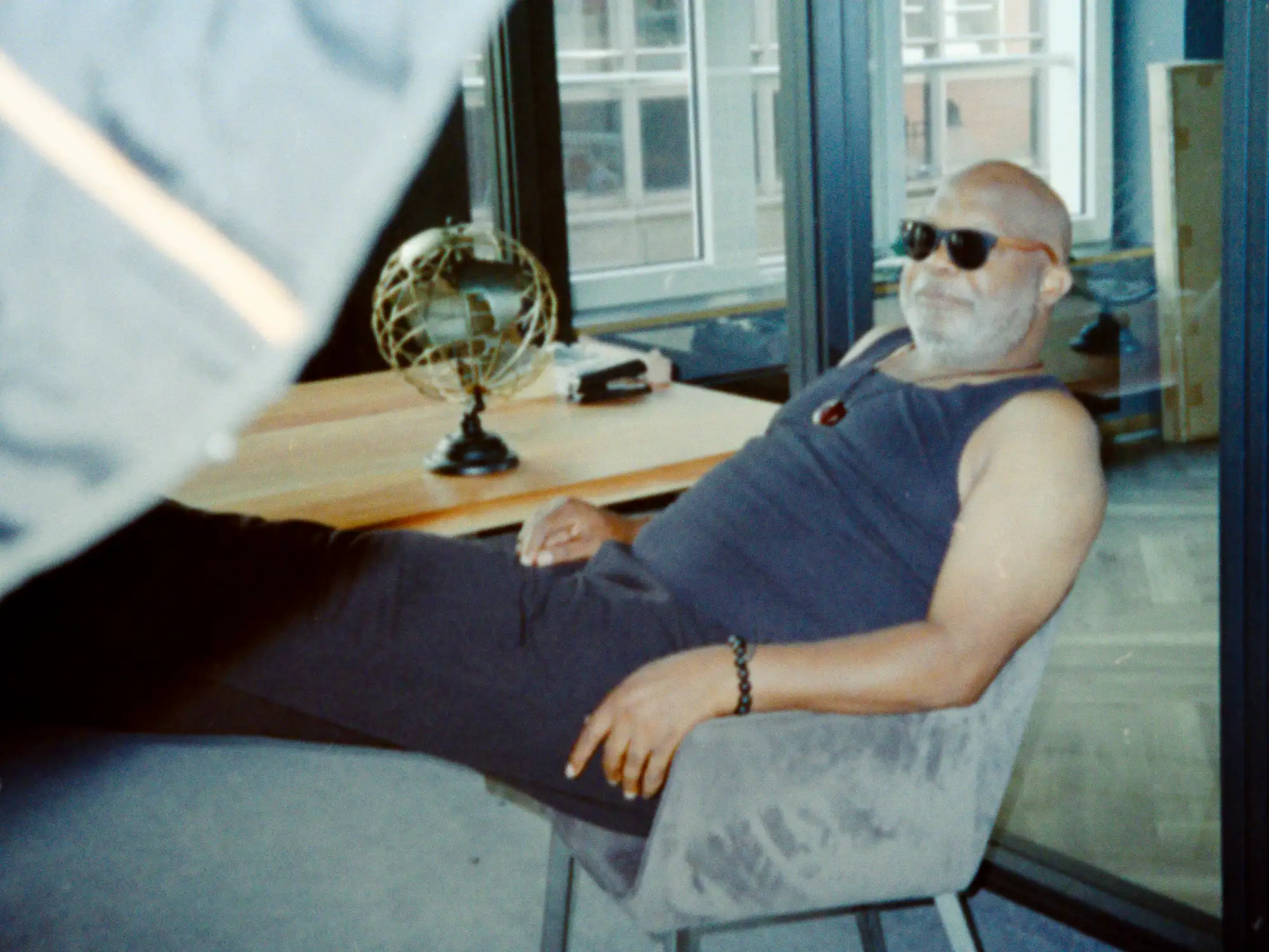 Behind the Scenes: Männliches Model mit Sonnenbrille sitzt entspannt auf einem Bürostuhl neben dem Schreibtisch.
