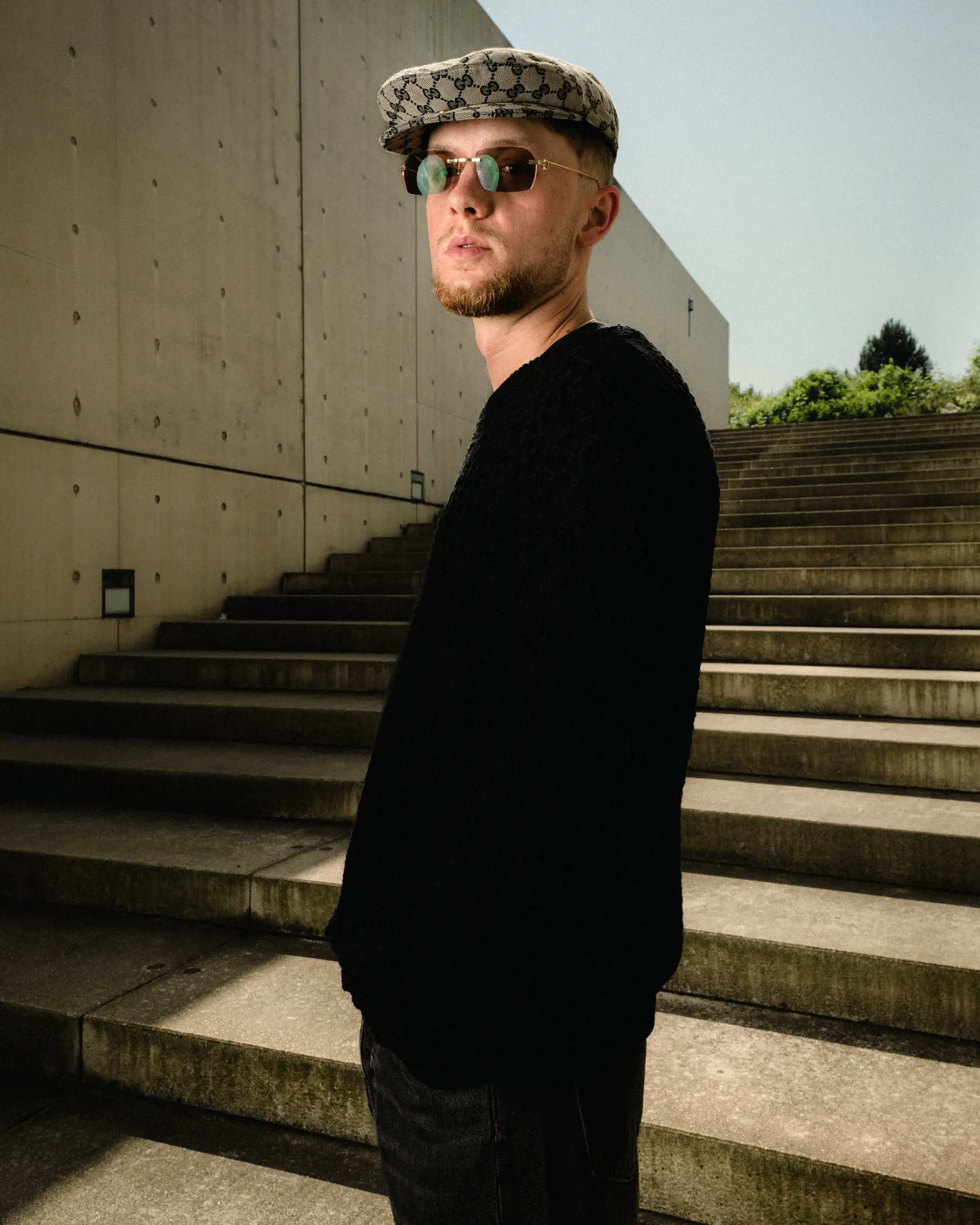 Strom im schwarzen Outfit mit Sonnenbrille und gemusterter Kappe neben einer Betontreppe.
