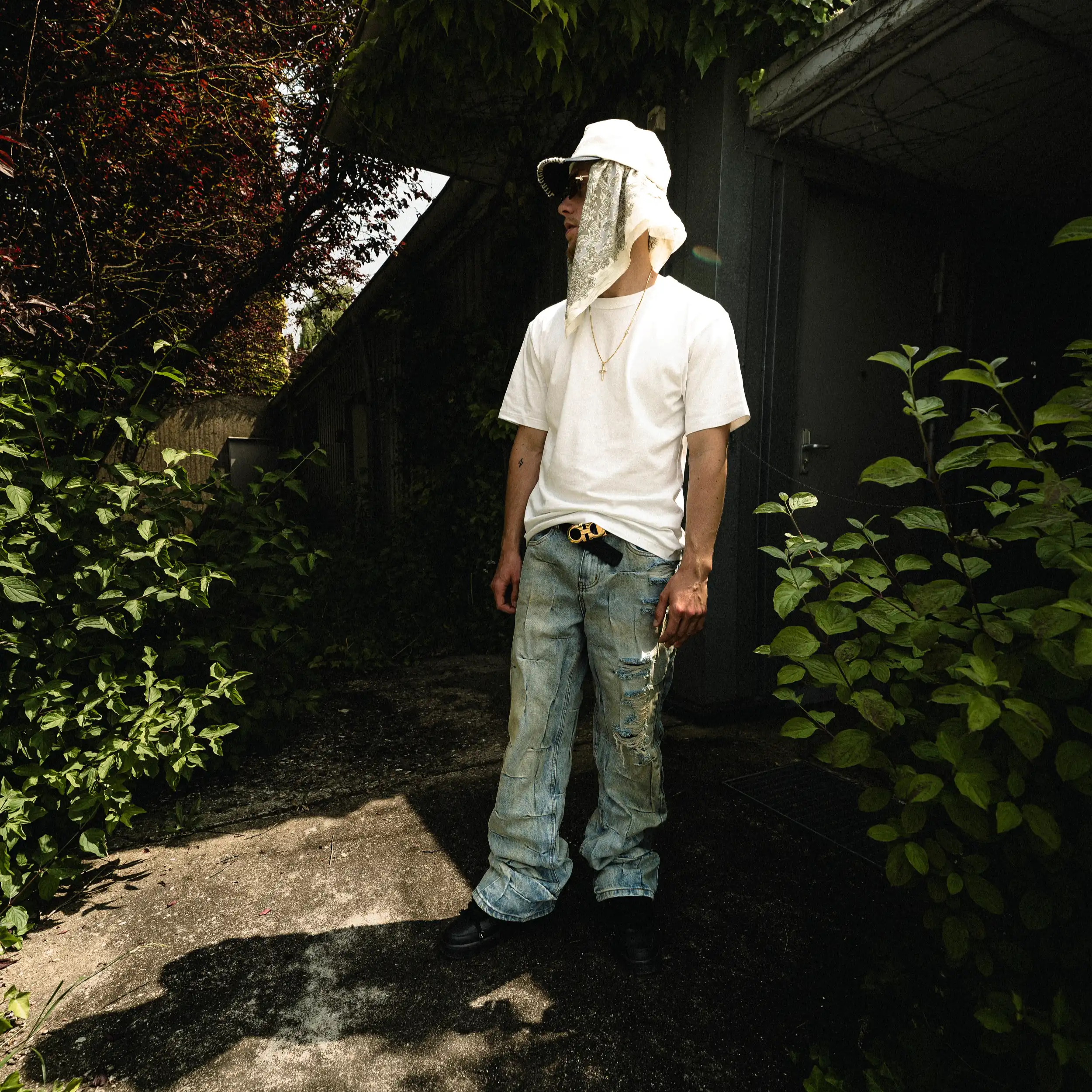 Strom im weißen T-Shirt und heller Jeans mit Kopftuch und Sonnenbrille auf einem Weg zwischen Büschen.