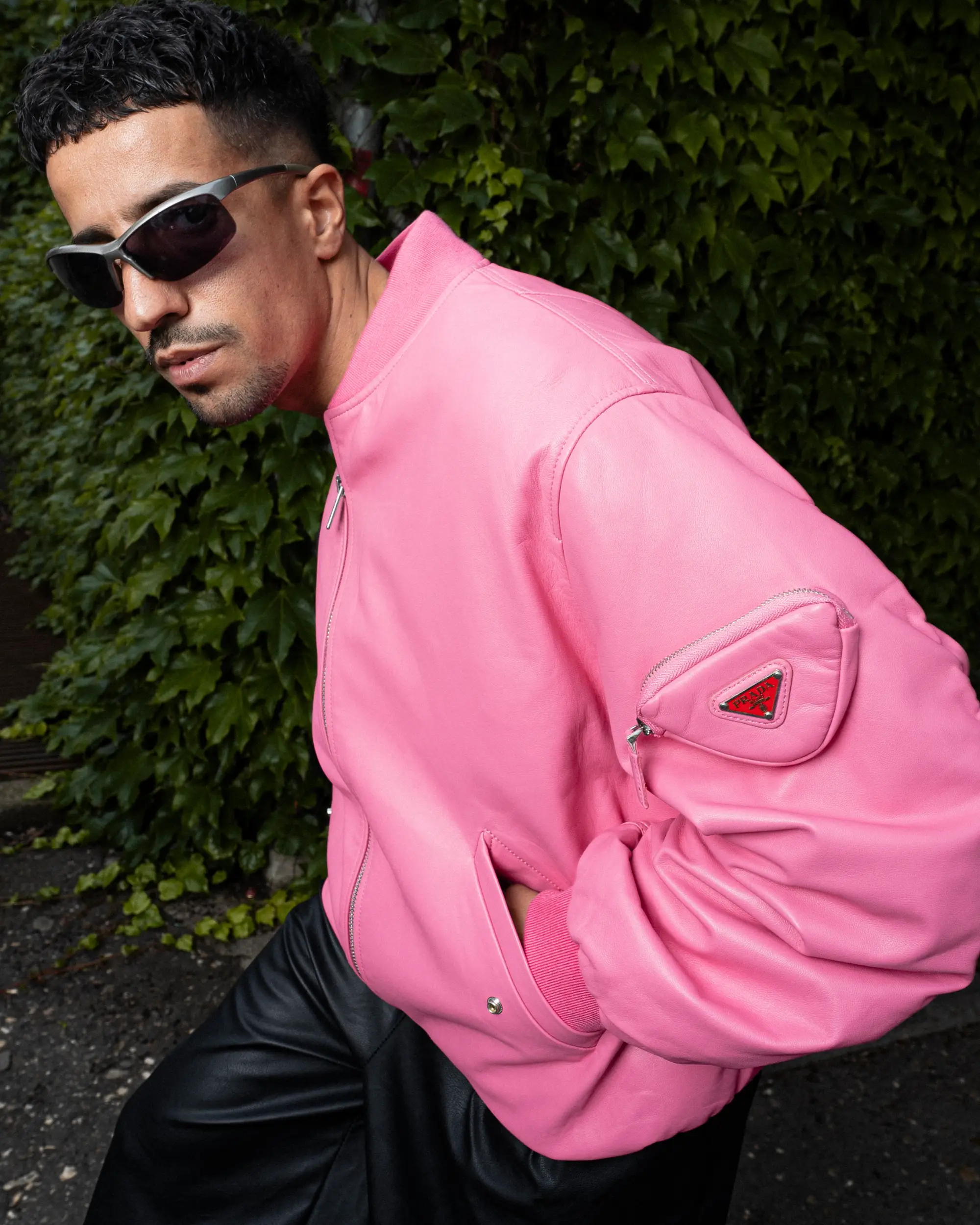 Ahmad mit Sonnenbrille in einer pinken Prada-Bomberjacke vor einer begrünten Wand beim Fashion-Shooting in Köln.
