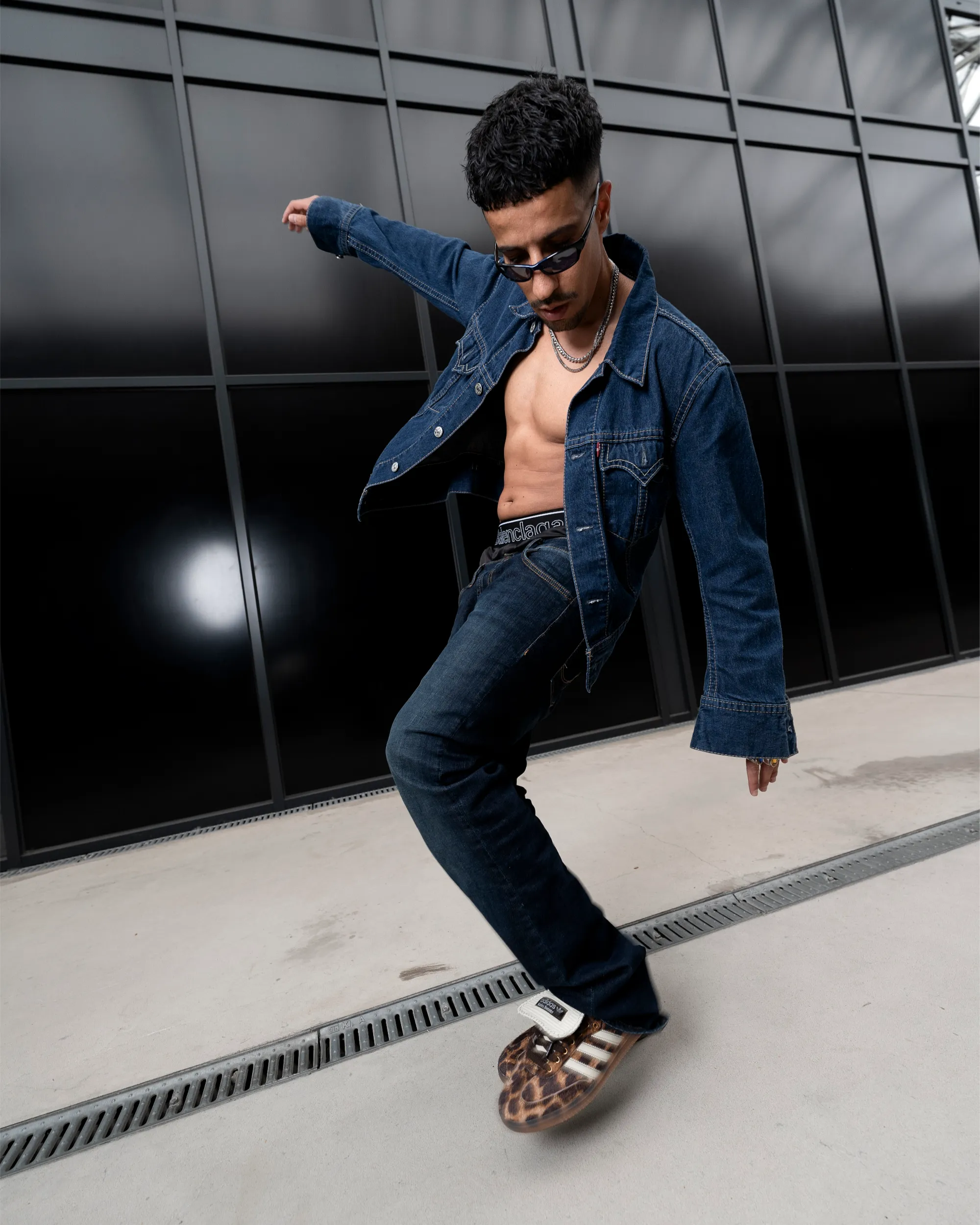 Ahmad im Denim-Look mit offener Jeansjacke und Sonnenbrille in einer Bewegungspose unter einem Glasdach.