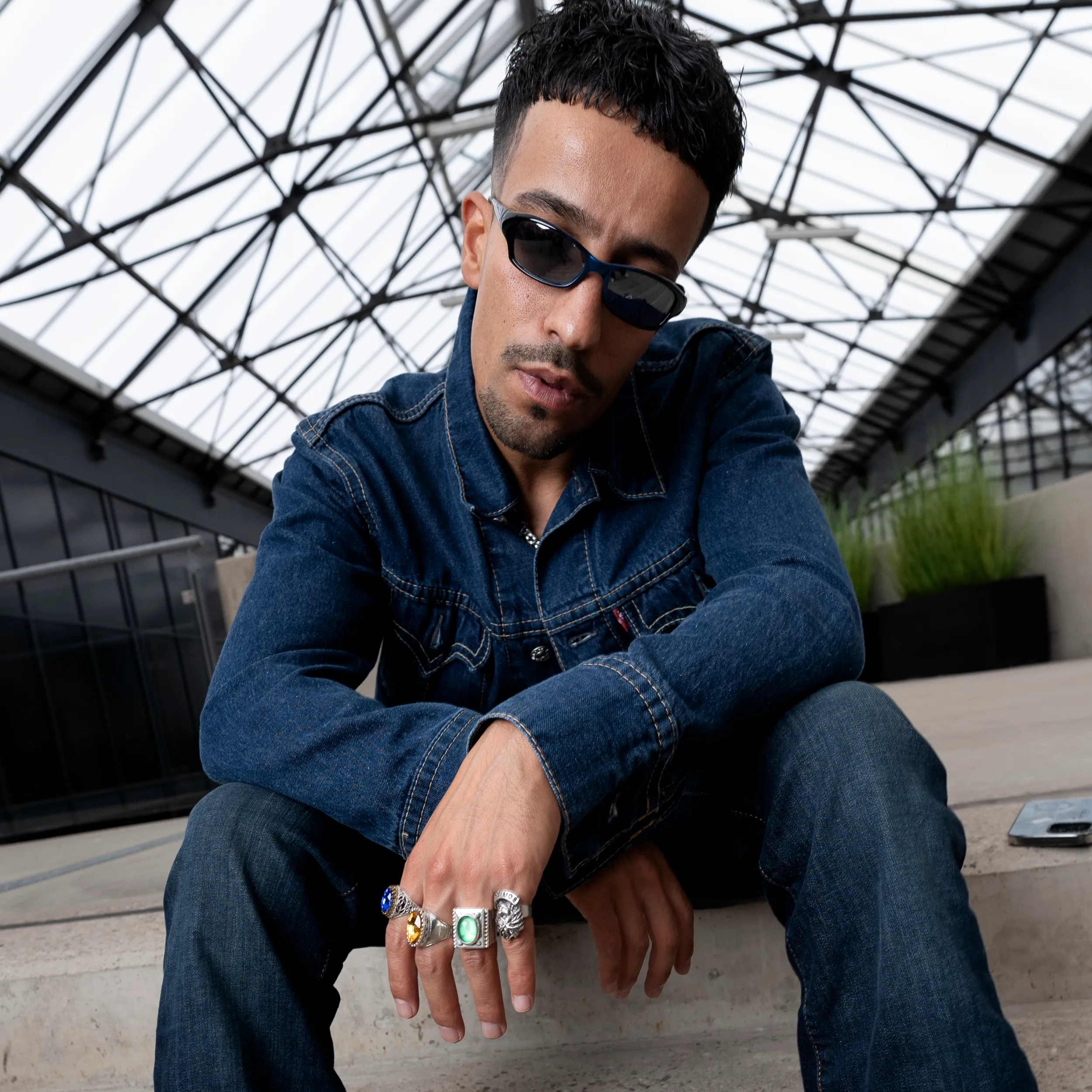 Ahmad sitzt im Denim-Look unter einem Glasdach und zeigt seine mit Ringen geschmückte Hand.