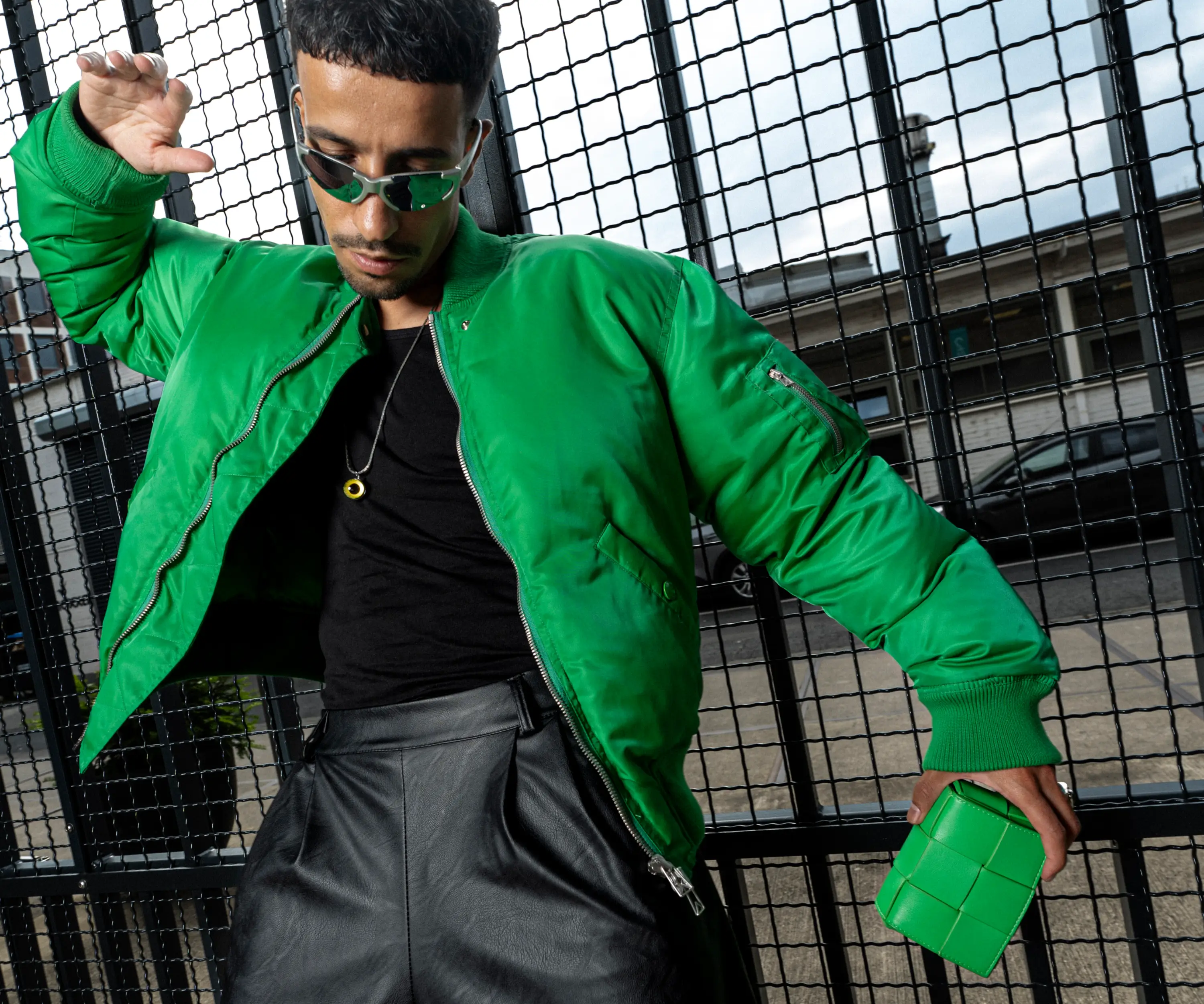 Ahmad mit Sonnenbrille, grüner Bomberjacke und schwarzem Outfit vor einem Metallgitter.