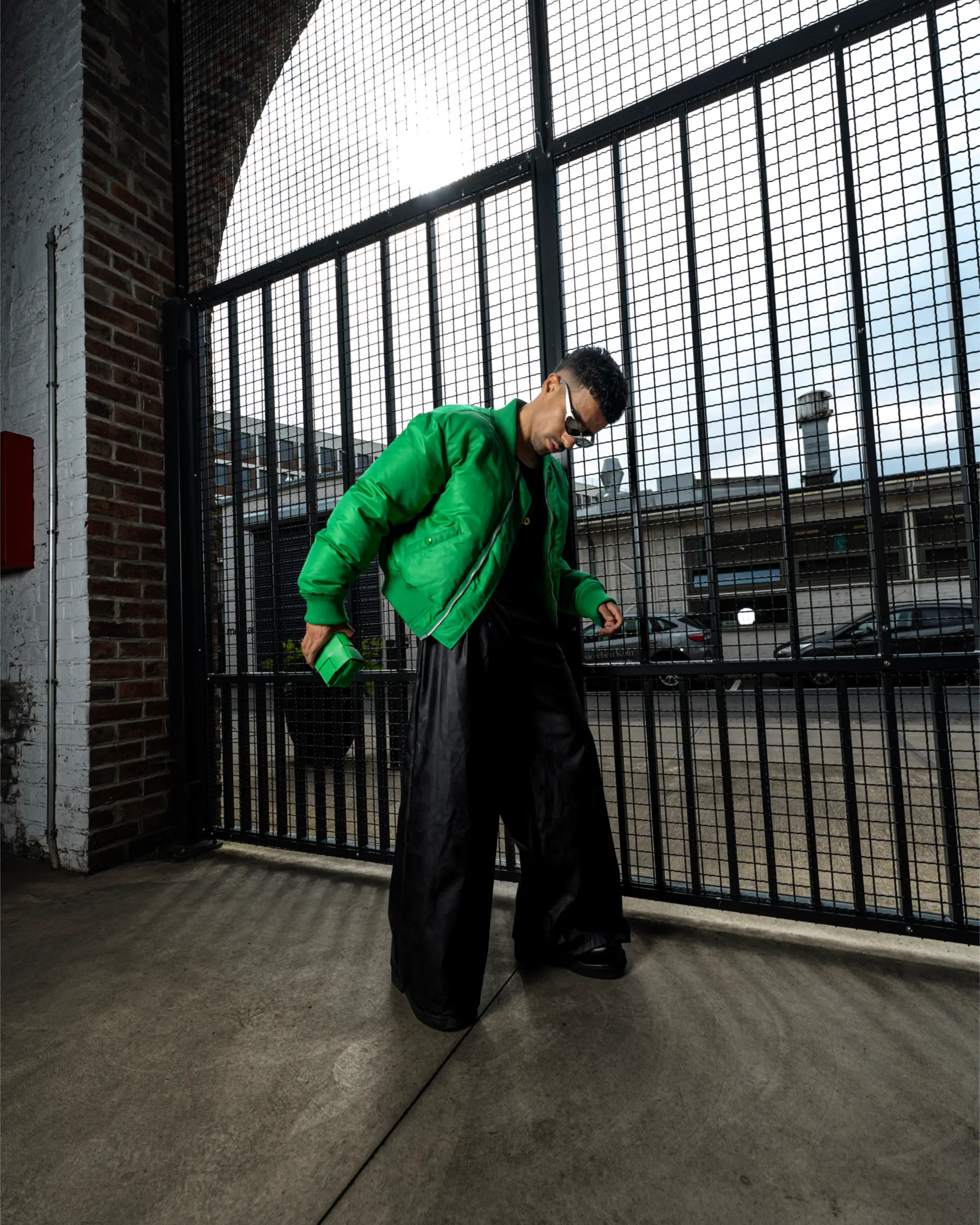 Ganzkörperaufnahme von Ahmad in grüner Bomberjacke und weiter schwarzer Hose vor einem hohen Metallgitter.