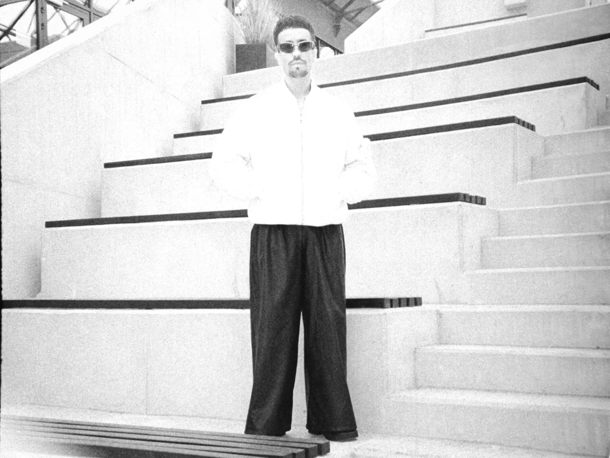 Analoge Schwarz-Weiß-Ganzkörperaufnahme von Ahmad im hellen Oberteil und weiter dunkler Hose vor einer Treppe.