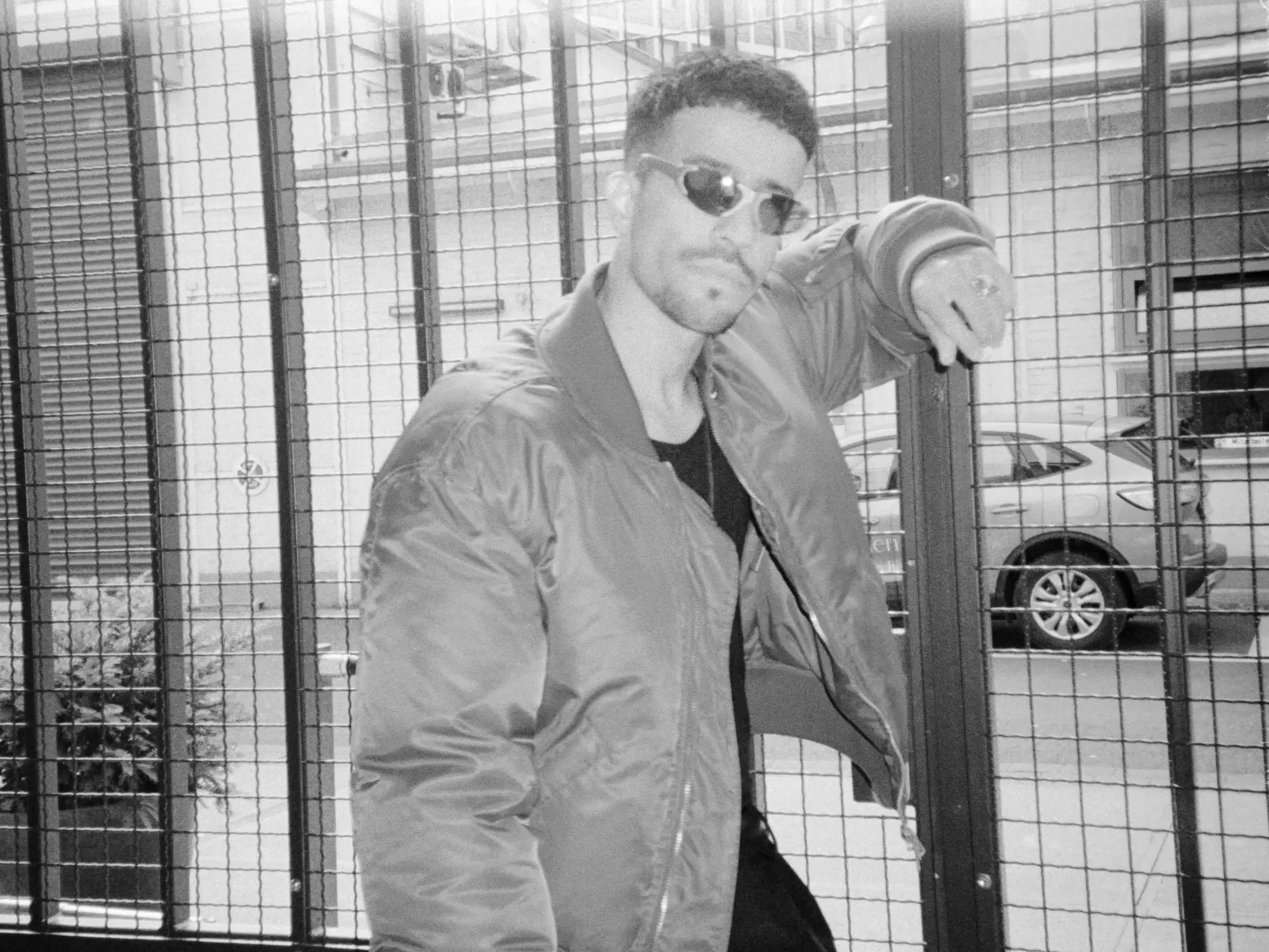 Analoges Schwarz-Weiß-Porträt von Ahmad mit Sonnenbrille und Bomberjacke vor einem Metallgitter in Köln.