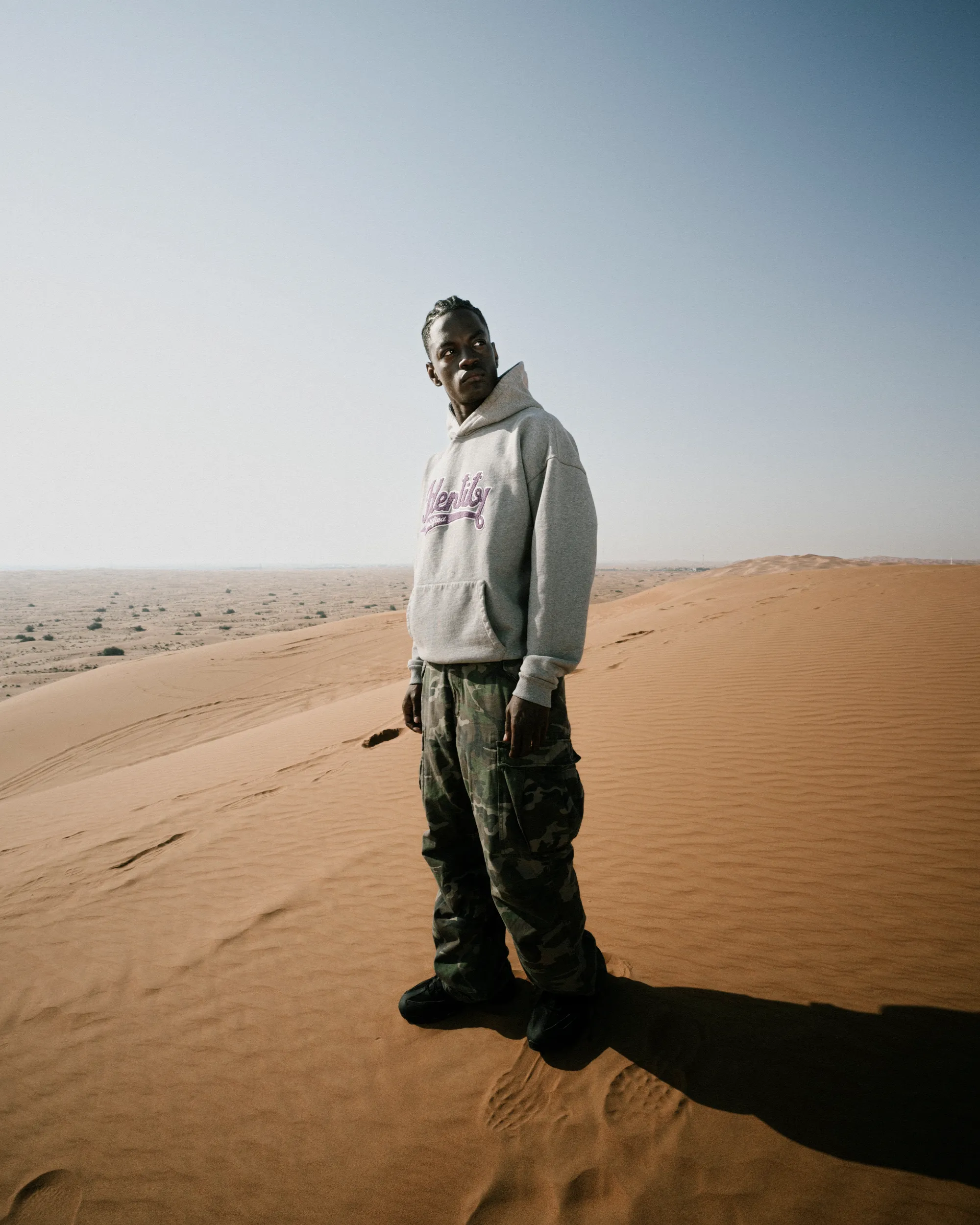 Ganzkörperaufnahme von Ibo Ouro-Bodi im grauen Hoodie und Camouflagehose auf einer Sanddüne.