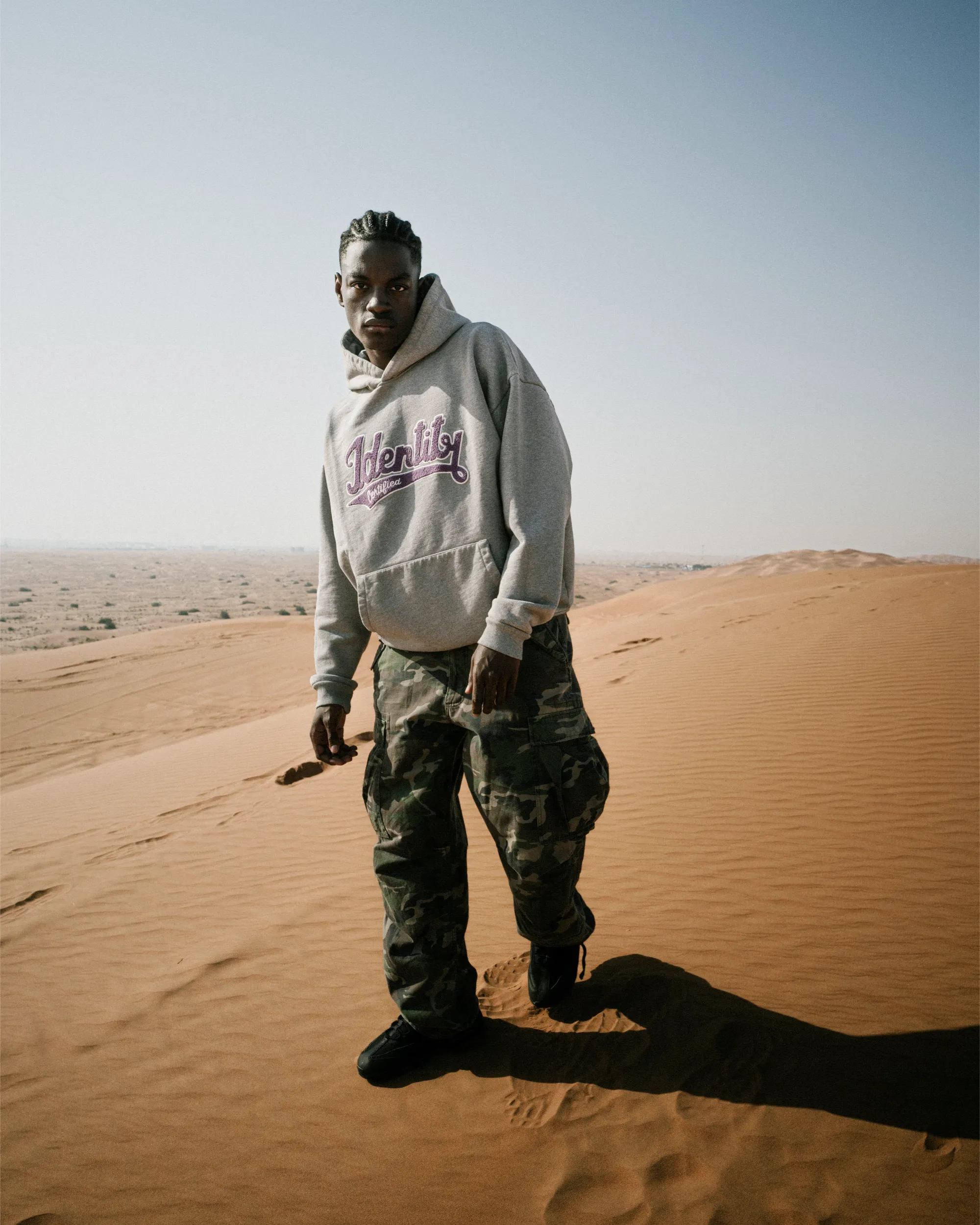Ibo Ouro-Bodi steht im grauen Hoodie und Camouflagehose auf einer Düne mit langem Schatten im Sand.
