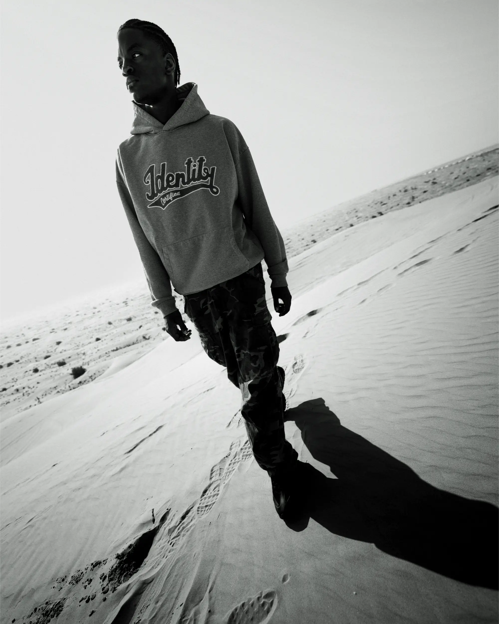 Schwarz-Weiß-Aufnahme von Ibo Ouro-Bodi im Hoodie auf einer Sanddüne mit markantem Schatten.