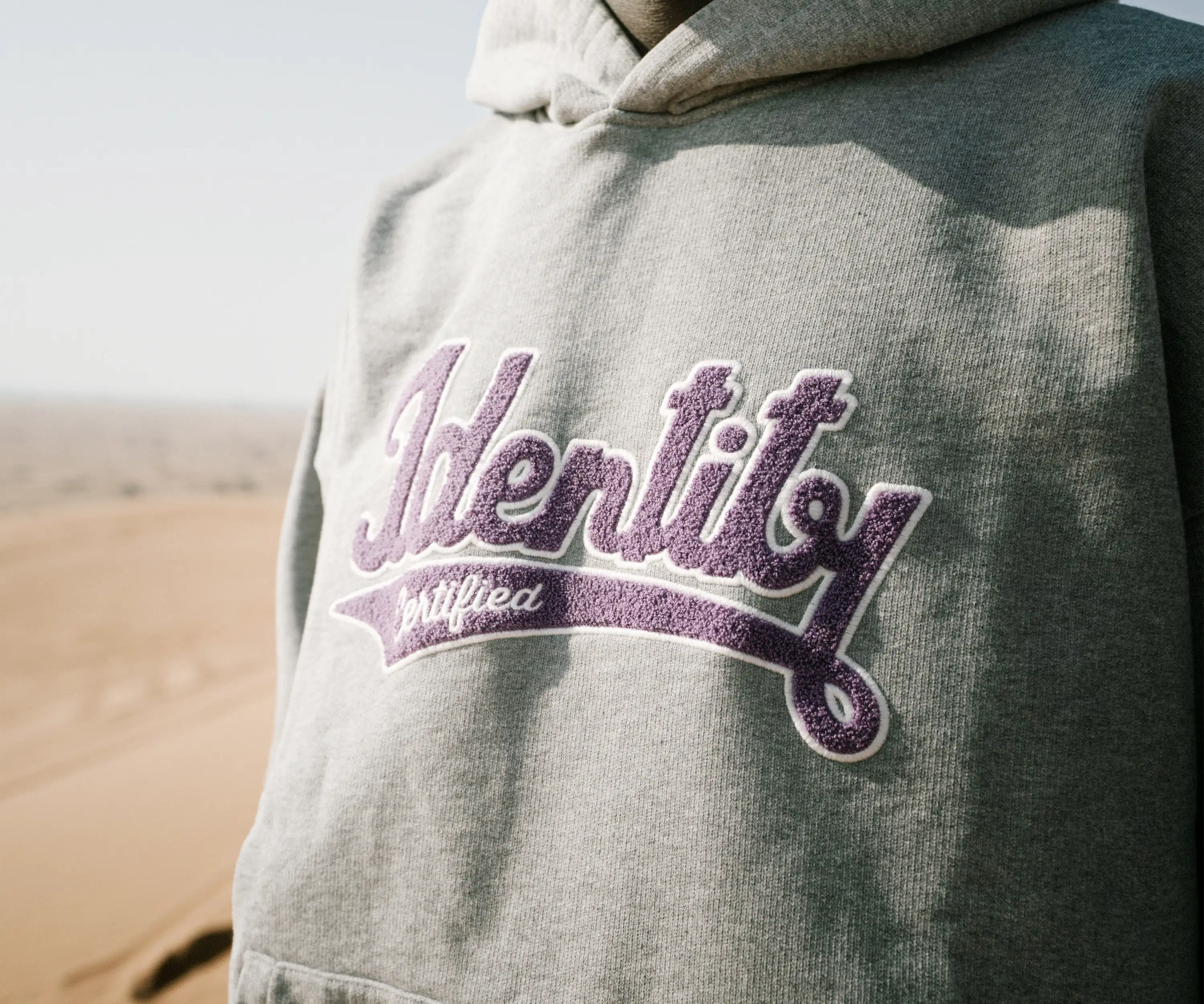 Detail der lilafarbenen Identity-Stickerei auf einem grauen Hoodie der Certified Identity Signature Collection.